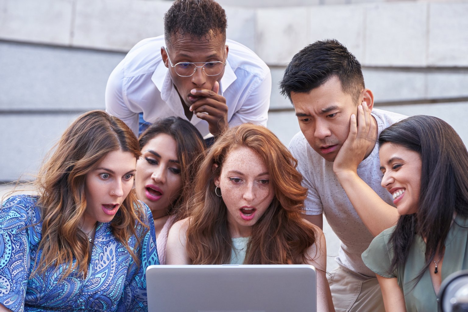Group of multi-ethnic people look at laptop screen with a shocked expression on their faces while sitting on stairs outdoors. Urban lifestyle, technology and friendship concept.