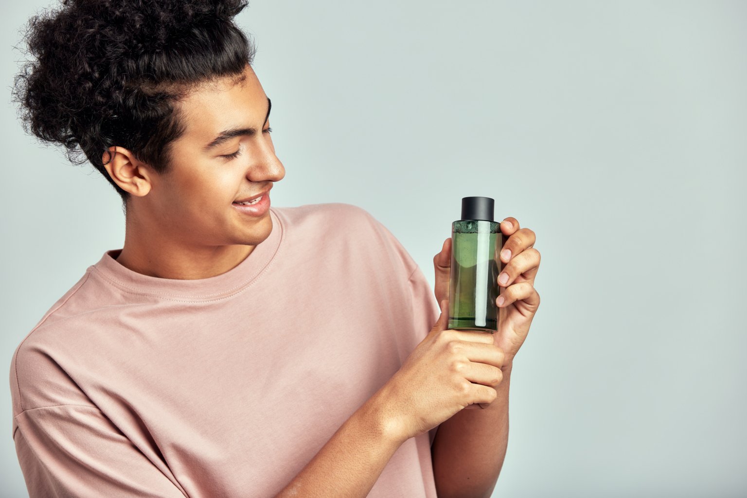 Portrait of young handsome smiling guy with a bottle of facial lotion in his hands. Swarthy man with black curly hair practice skincare routine to maintain his youthful and healthy appearance