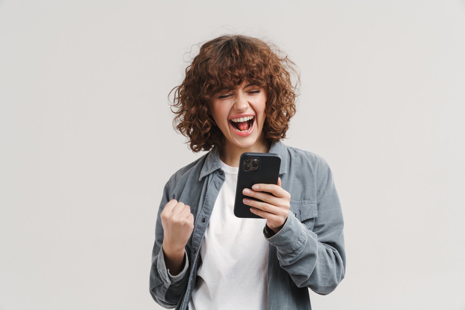 Young excited woman making winner gesture while using mobile phone isolated over white background