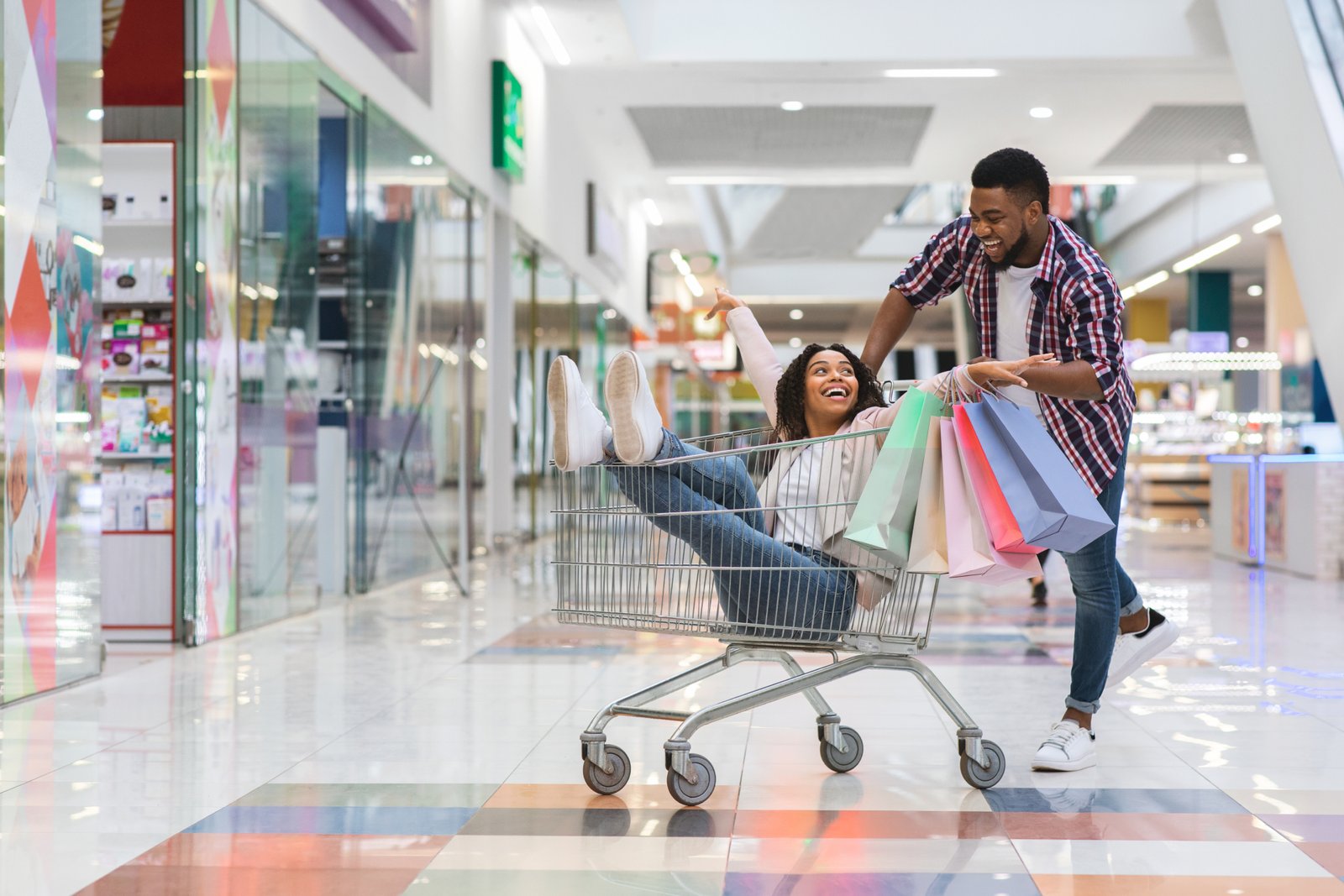 Cheerful black man pulling shopping trolley with his girlfriend inside, having fun while making purchases together in modern department store, fooling and laughing, enjoying seasonal sales, copy space