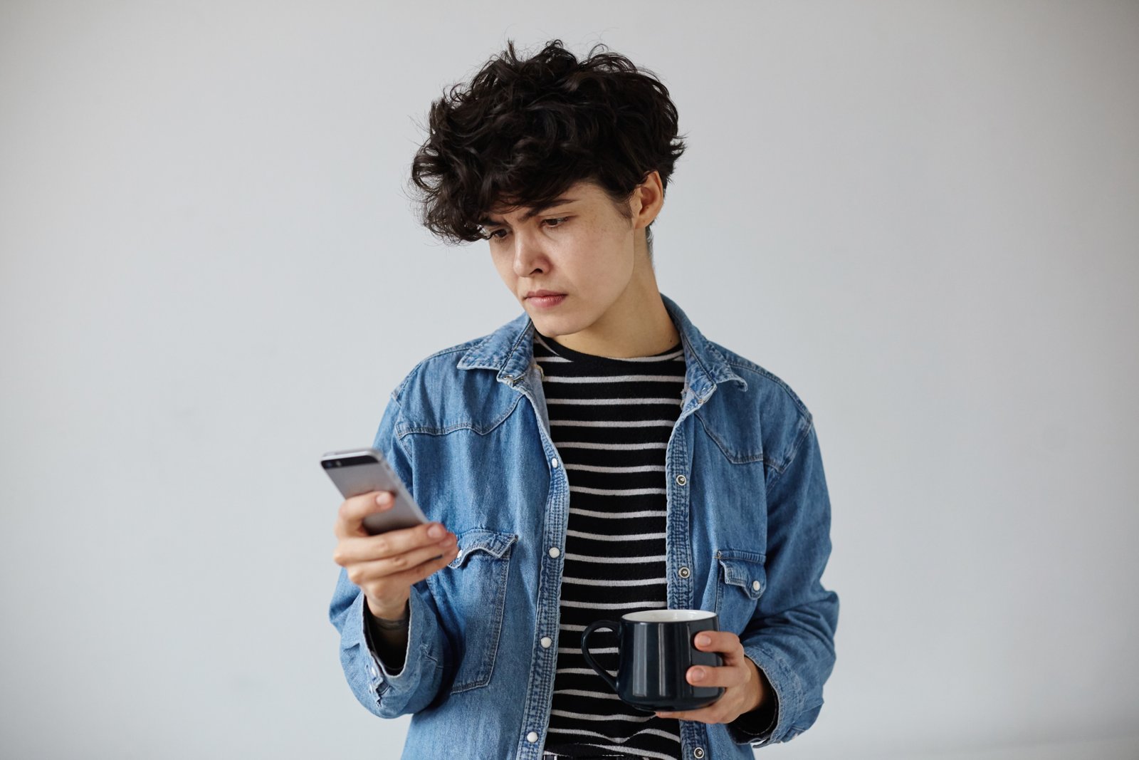 Puzzled young pretty curly brunette female with short trendy haircut holding smartphone in her hand and looking confusedly at screen, posing over white background with dark ceramic cup