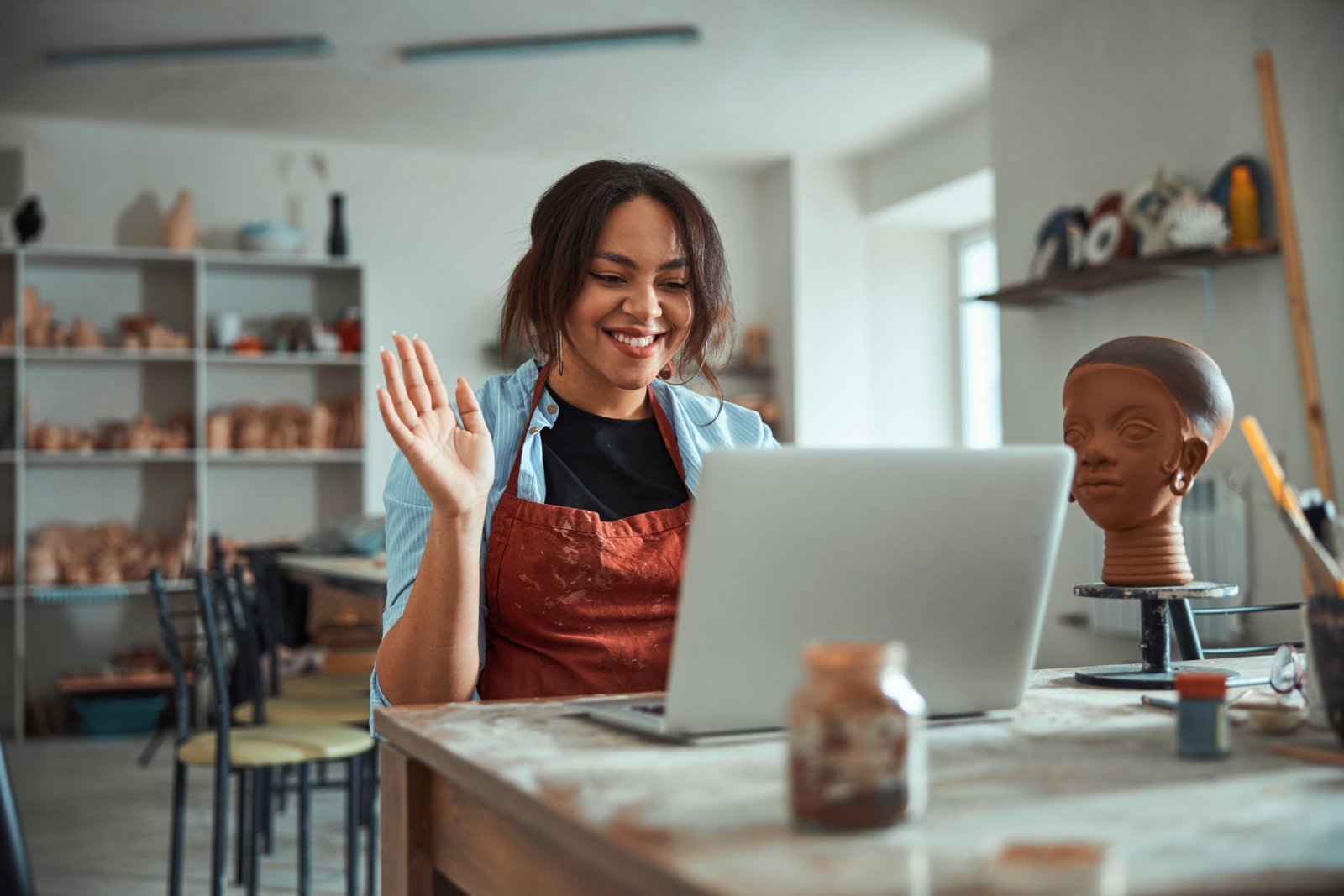 Beautiful young woman ceramic artist doing hello gesture and smiling while talking with friend through video call
