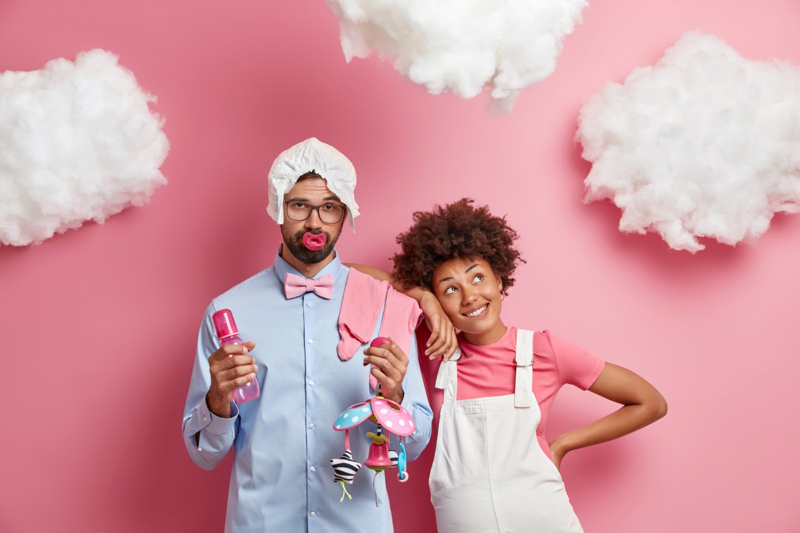 Happy mixed race parents expect baby prepare to become father and mother. Pleased Afro American pregnant woman leans at shoulder of husband who holds feeding bottle and toy. Expectation concept